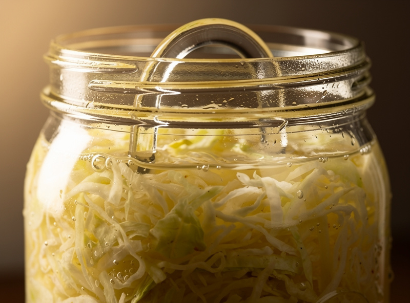 Macro close-up of a glass fermentation weight inside a wide-mouth mason jar holding sauerkraut submerged below the brine line, with the weight's finger-grip handle visible above the brine