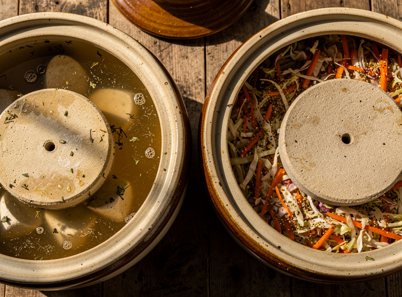 Top-down view of two large stoneware fermentation crocks side by side with lids removed showing ceramic weights inside, one half submerged in brine and the other on top of fermented vegetables