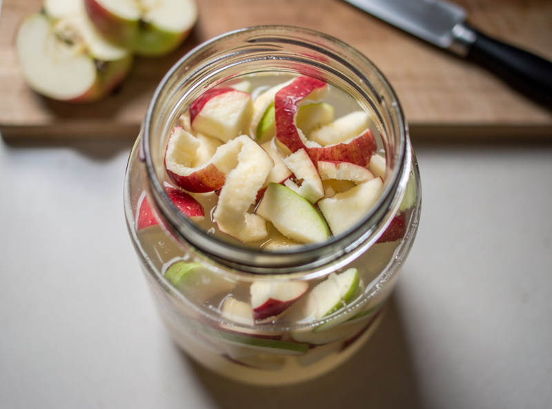 Top-down photograph of chopped apple cores, peels, and apple chunks in a wide-mouth glass jar partially submerged in sugar water