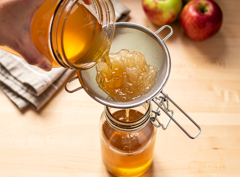 Hand pouring finished apple cider vinegar through a fine mesh strainer into a clean glass swing-top bottle with vinegar mother visible