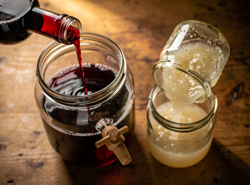 Top-down view of a half-empty bottle of red wine being poured into a glass jar with a wooden spigot alongside a small jar containing a vinegar mother culture being added to start fermentation
