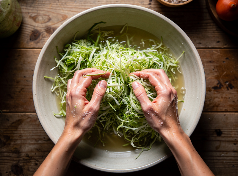 Hands massaging salted shredded green cabbage as brine pools in the bowl