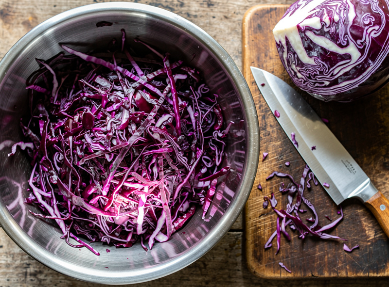 Finely shredded raw purple red cabbage in a stainless steel bowl beside a whole head and a knife