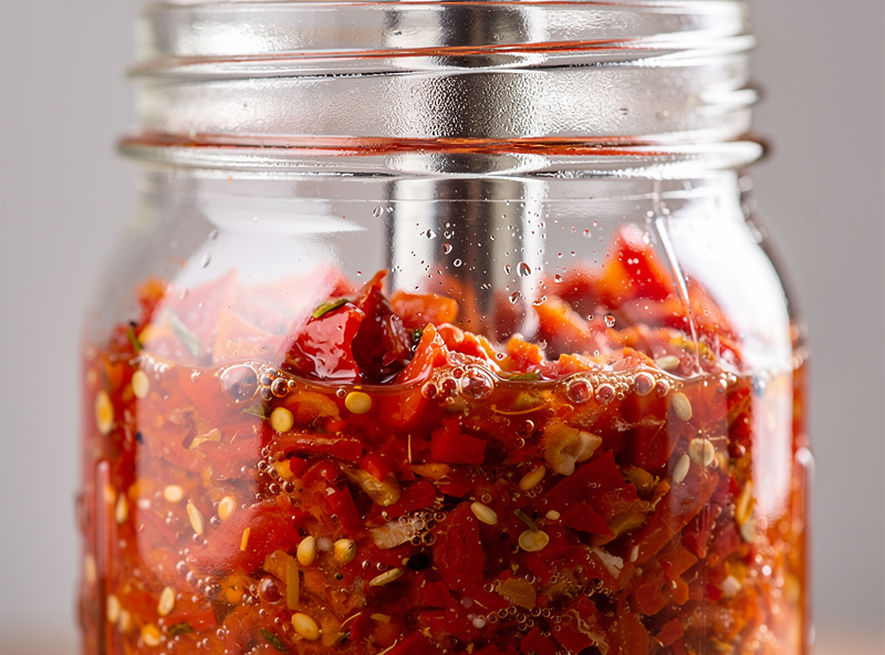 A jar packed with coarse red pepper mash fermenting in its own salt-drawn liquid under a glass weight, with no added brine