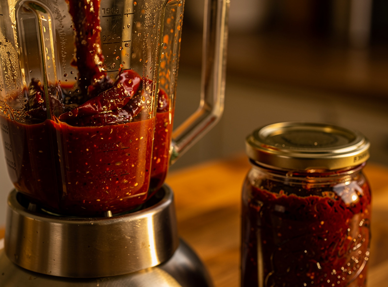 Finished dark red pepper mash being blended into a thick concentrated hot sauce, with a jar of fermented mash beside the blender
