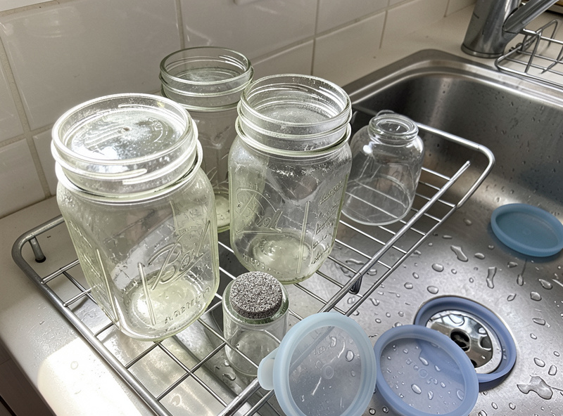 Clean mason jars, glass weights, and silicone lids drying upside down on a rack beside a sink