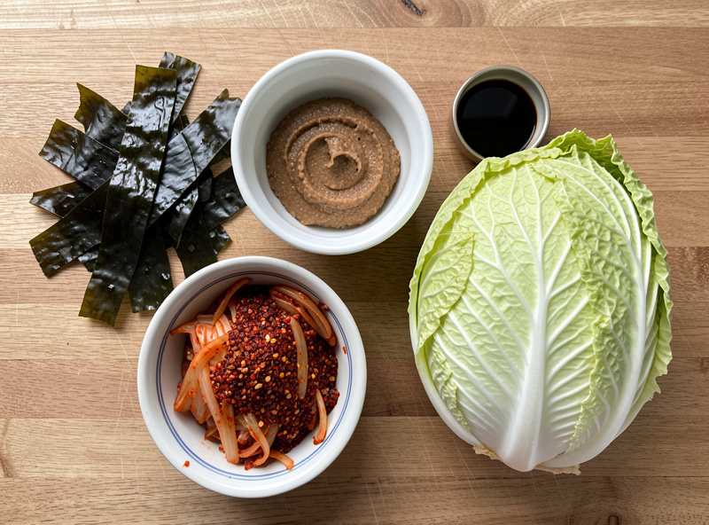 Vegan kimchi ingredients laid out on a counter including dried kombu kelp, dried shiitake mushrooms, miso paste, soy sauce, gochugaru, and napa cabbage