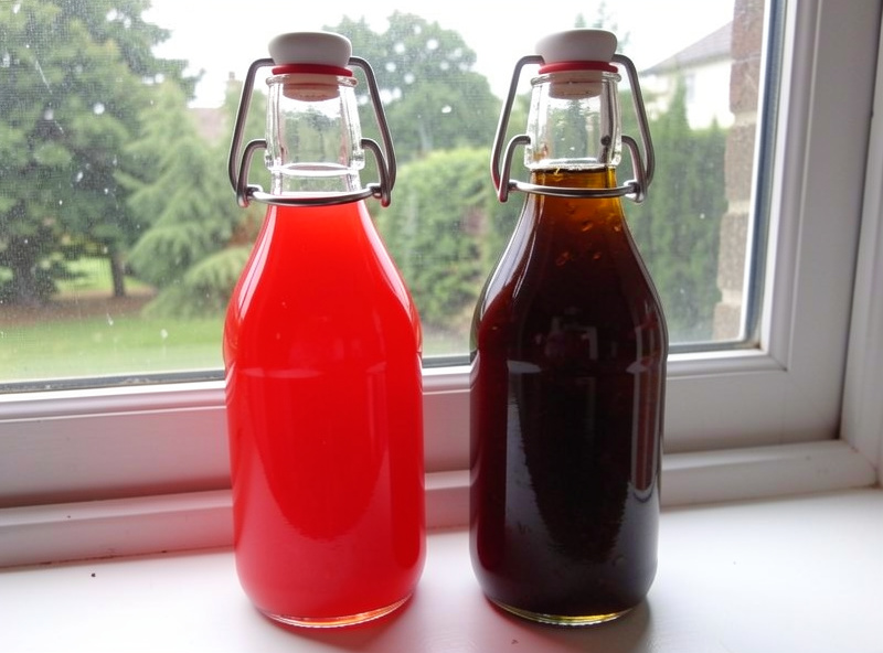 Two finished swing-top bottles of shrub syrup, one bright fresh red and one darker jammy amber, labeled, on a windowsill