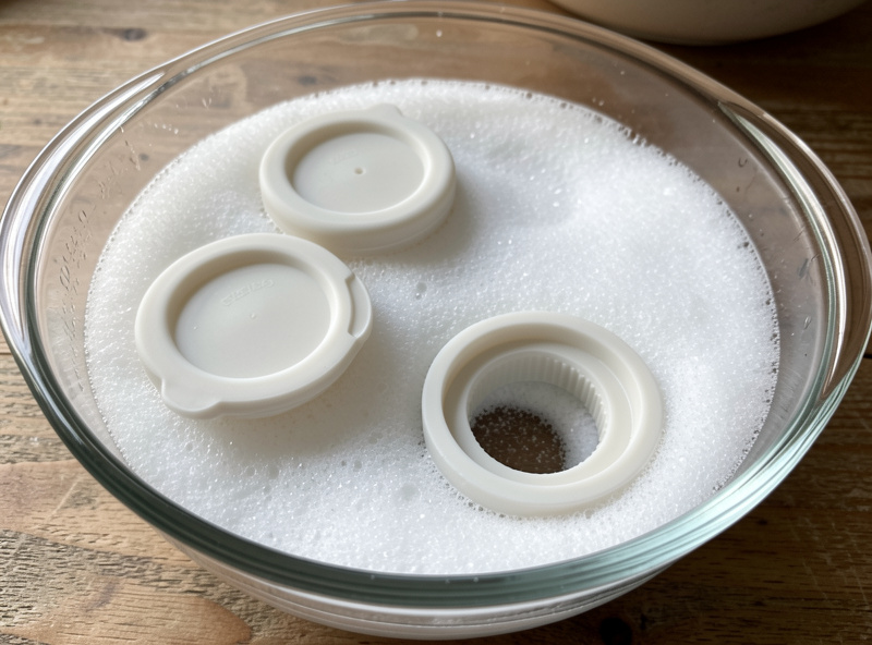 Silicone airlock lids and jar gaskets soaking in a glass bowl of cloudy baking-soda solution